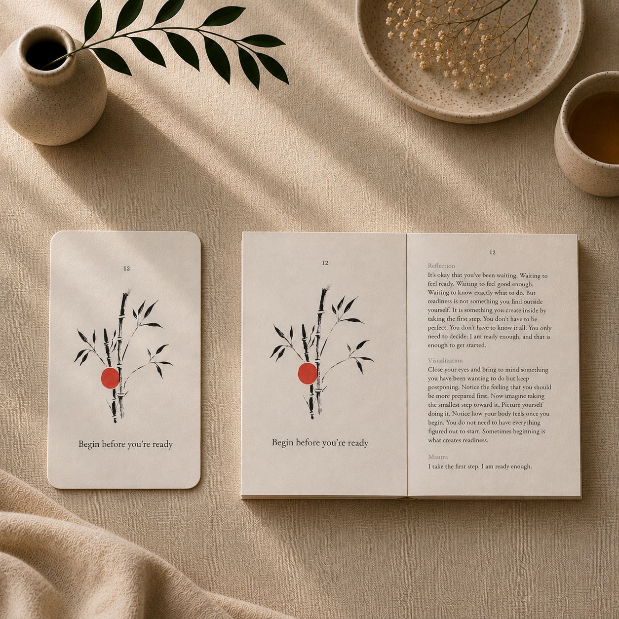 A styled flat-lay of a card and an open book or journal on beige fabric. Both pieces feature minimalist black ink bamboo illustrations with a small red circle and the text 'Begin before you're ready.
