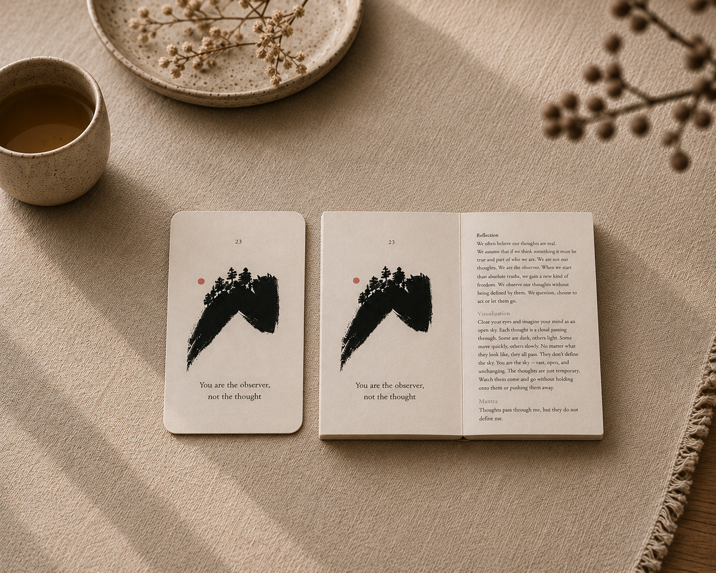 An open card deck book and two cards displayed on a beige fabric surface, styled with a cup of tea, dried flowers, and a ceramic dish in soft natural light.