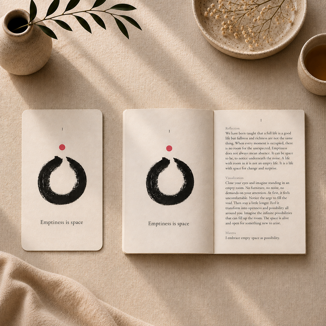 An open meditation card deck or journal spread laid on a beige fabric surface, showing minimalist black circular artwork with a small red dot and the text 'Emptiness is space' on both cards. The scene includes a small vase with greenery, dried flowers, and a cup in a styled flat-lay setup.