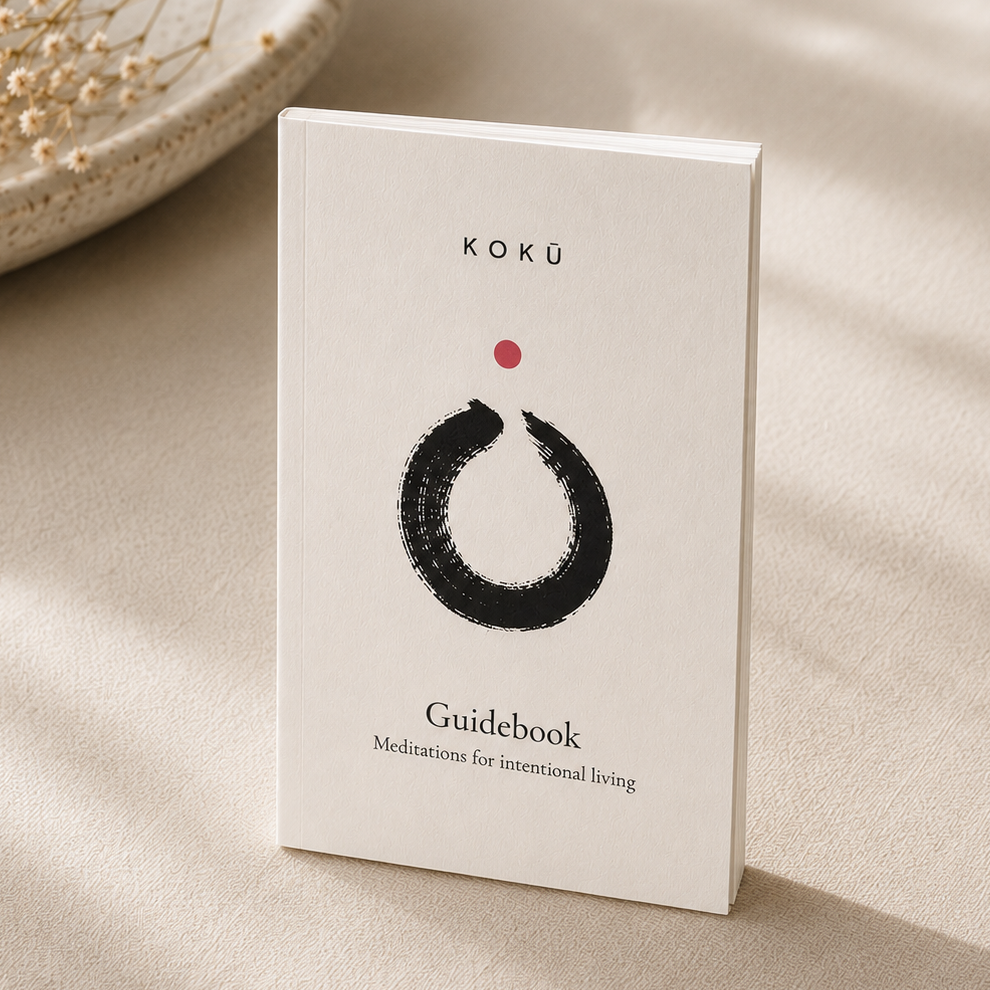 A white guidebook titled 'KOKÜ Guidebook' with a minimalist black brushstroke circle and a small red dot on the cover, standing upright on a beige fabric surface with soft shadows and dried flowers in the background.
