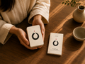 A person in a beige robe holds a small white set of meditation cards with a black circular logo and red dot, while another copy rests on a wooden table. A ceramic vase and bowl are visible in the warm sunlight.
