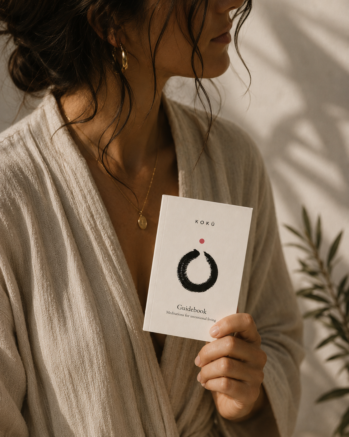 A woman in a beige linen robe holds a white card titled 'KOKU Guidebook' with a minimalist black and red circular logo. The background features warm, natural light shadows and a leafy plant.
