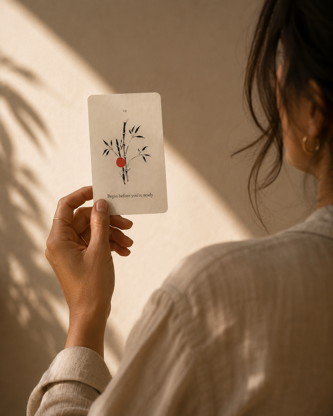 A person in a beige linen shirt holds up a white oracle card featuring a minimalist black ink illustration of bamboo with a small red fruit and the text 'Begin before you study'. The background is a warm beige wall with soft leaf shadows.