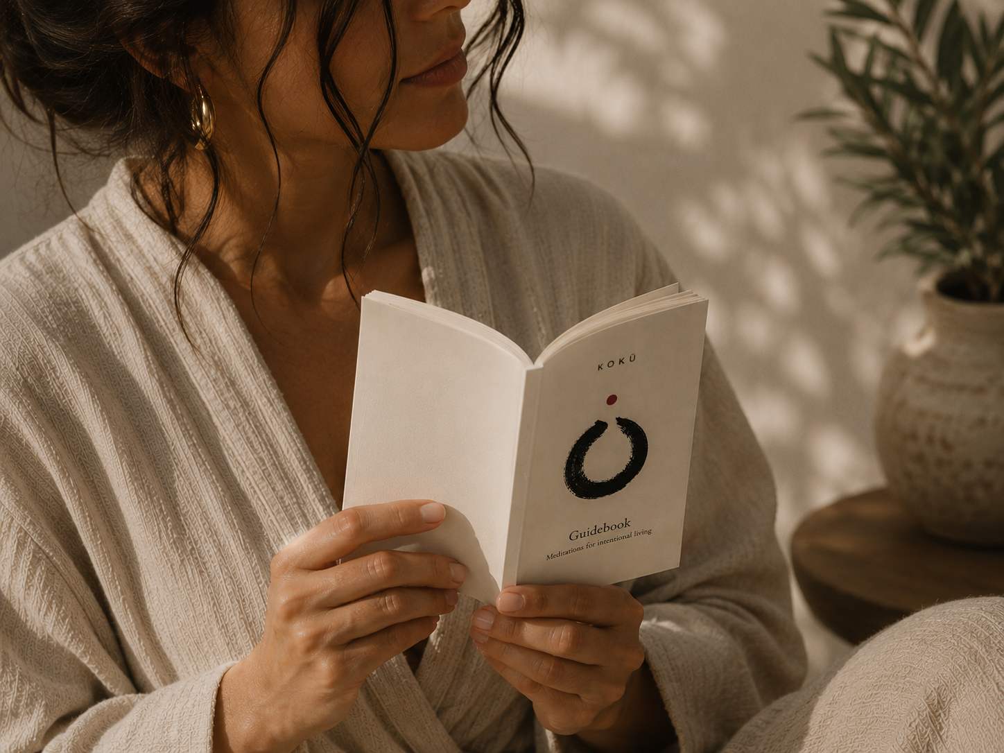 A woman in a beige robe holds an open minimalist guidebook with a black circular logo and the title 'Guidebook' on the cover, set in a softly lit indoor environment with a potted plant in the background.